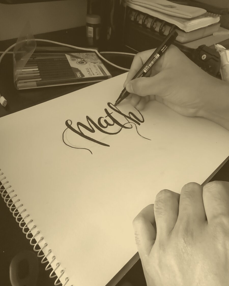 A hand lettering the word Math in brush pen on a spiral pad.